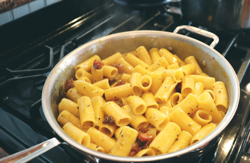 Carbonara pasta being mixed in an all clad silver pot on top of a black stove. 