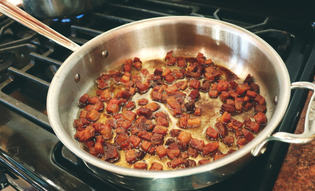 An all clad silver pan placed on a burner with guanciale meat cooking in it. 